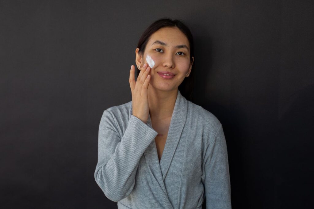Positive ethnic female in bathrobe applying moisturizing cream on cheek during daily beauty routine against black background and looking at camera