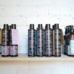 A variety of natural cosmetic bottles lined up on a wooden shelf against a white brick wall, suggesting eco-friendly beauty