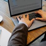 Crop faceless male entrepreneur in formal wear sitting at table with cup of coffee and notebook while working on project in laptop