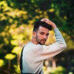 Side view of queer cool male in pullover touching hair while looking at camera on blurred background of trees
