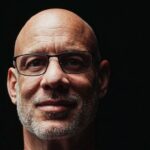 Close-up portrait of a bald man with glasses on a dark background