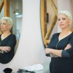 A professional hairdresser confidently stands in a modern salon with crossed arms, reflecting in a mirror