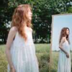 A woman in a white dress gazes at her reflection in a mirror amidst a serene outdoor setting