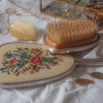 Elegant vintage vanity set with floral hand mirror, brush, and jewelry on a textured fabric