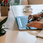 High angle side view of crop unrecognizable guy with smartwatch working remotely on laptop at table near cup of coffee on saucer near green potted plant and notebook in room