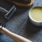 A flat lay of grooming tools including a razor, brush, and pomade on a textured surface